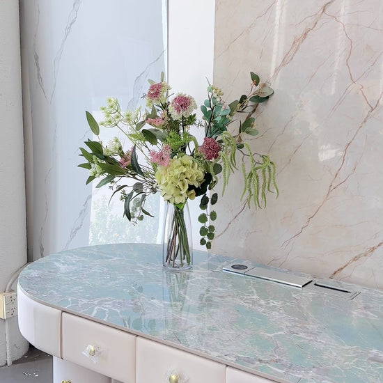 Artificial floral arrangement featuring green hydrangeas, pink wildflowers, and lush foliage in a clear glass vase with acrylic water