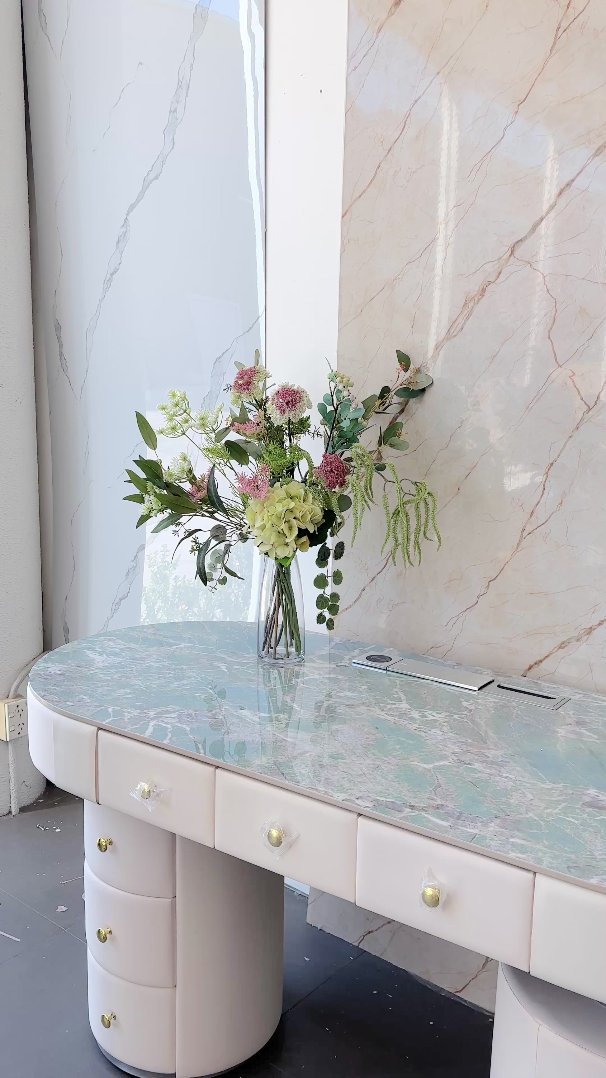 Artificial floral arrangement featuring green hydrangeas, pink wildflowers, and lush foliage in a clear glass vase with acrylic water