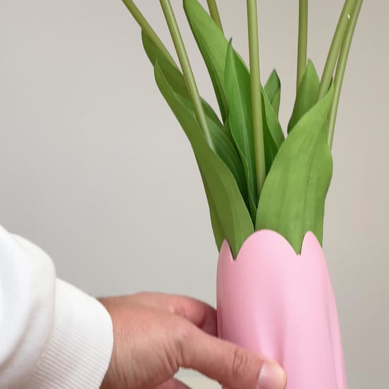 Artificial pink tulip arrangement with lifelike green stems and leaves, displayed in a matching pink vase with a scalloped tulip-shaped rim, set against a neutral background.