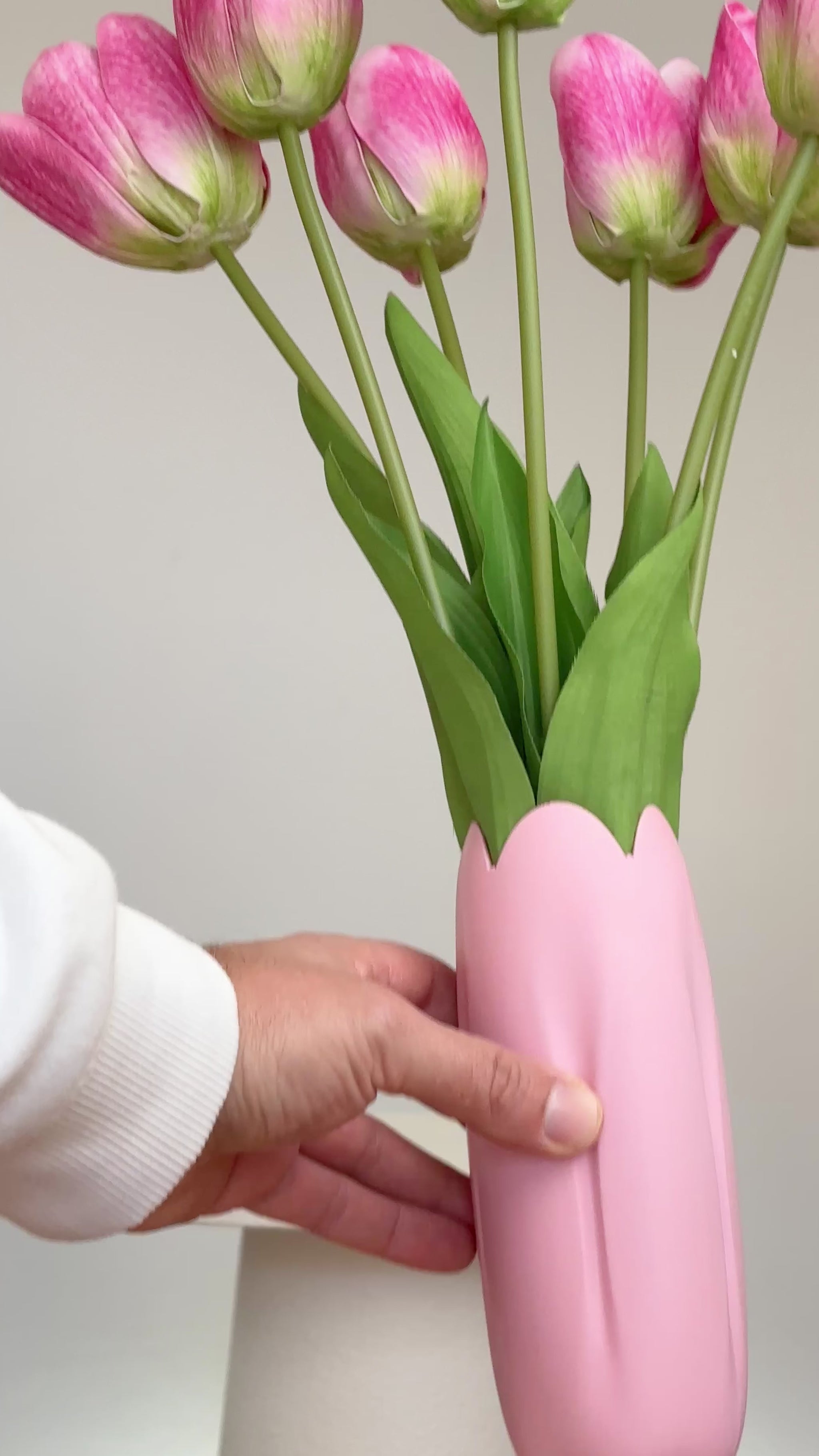 Artificial pink tulip arrangement with lifelike green stems and leaves, displayed in a matching pink vase with a scalloped tulip-shaped rim, set against a neutral background.