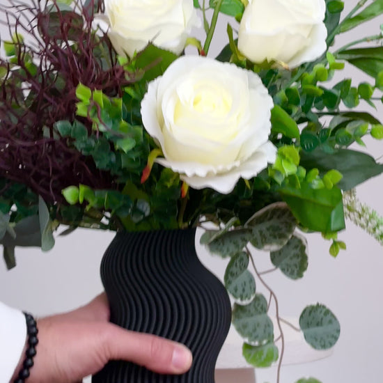 Artificial white rose arrangement with lush greenery in a sculptural matte black wavy vase, styled on a marble table — a modern and elegant decorative floral display ideal for minimalist interiors.
