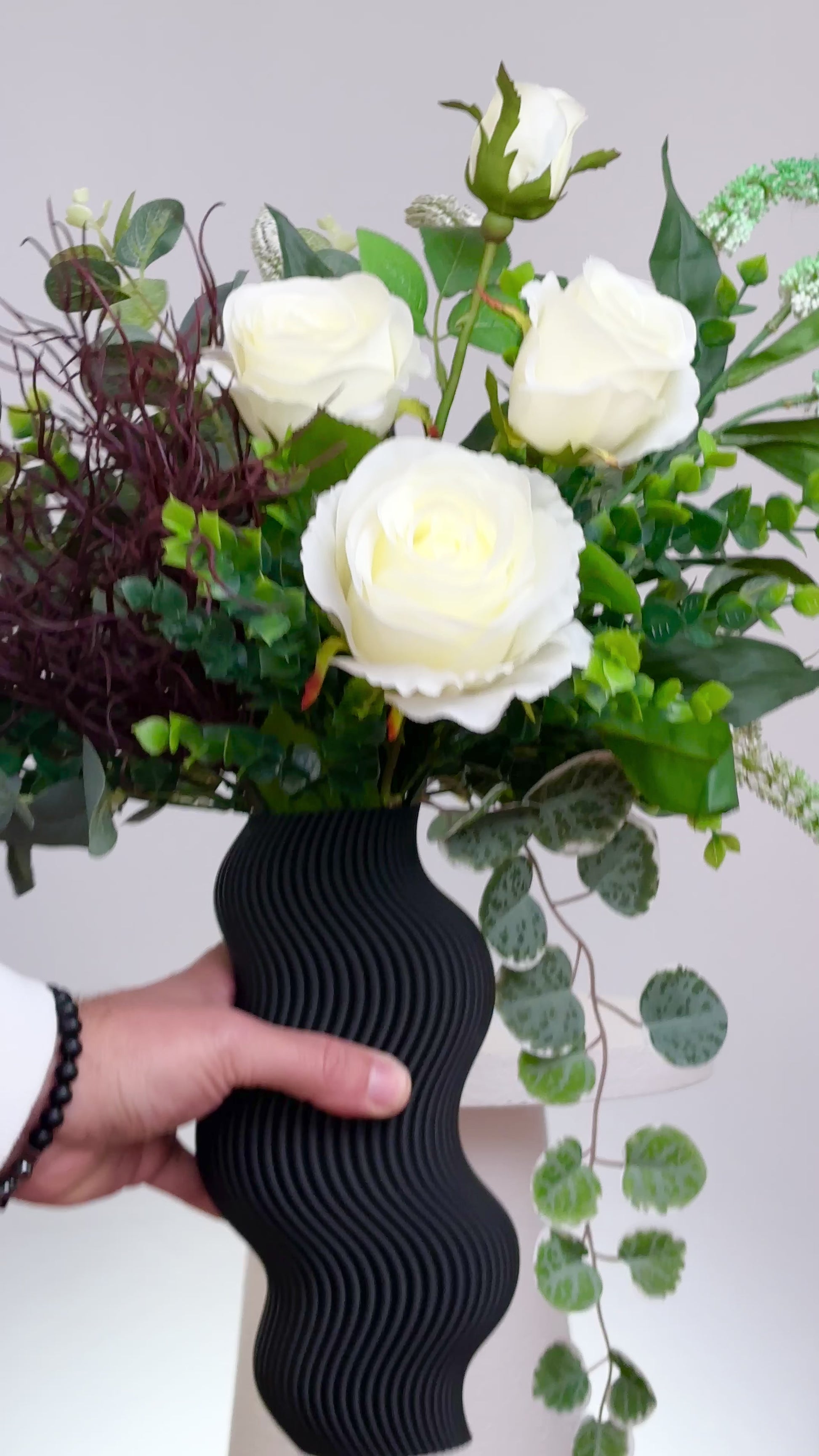 Artificial white rose arrangement with lush greenery in a sculptural matte black wavy vase, styled on a marble table — a modern and elegant decorative floral display ideal for minimalist interiors.