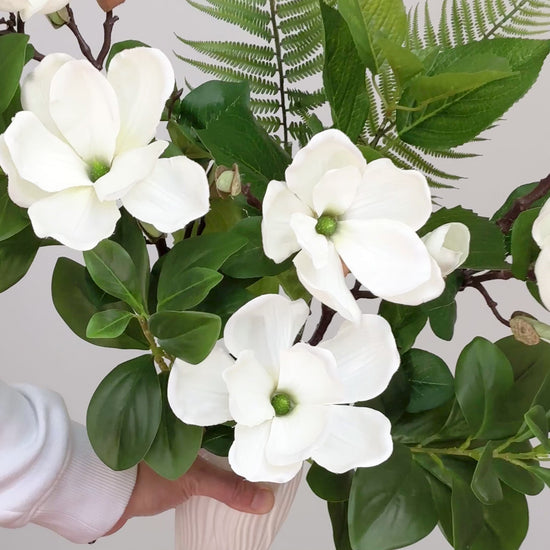 Artificial white magnolia flower arrangement with lush green leaves and fern accents, beautifully styled in a sleek white vase on a round white table, set against a neutral background.