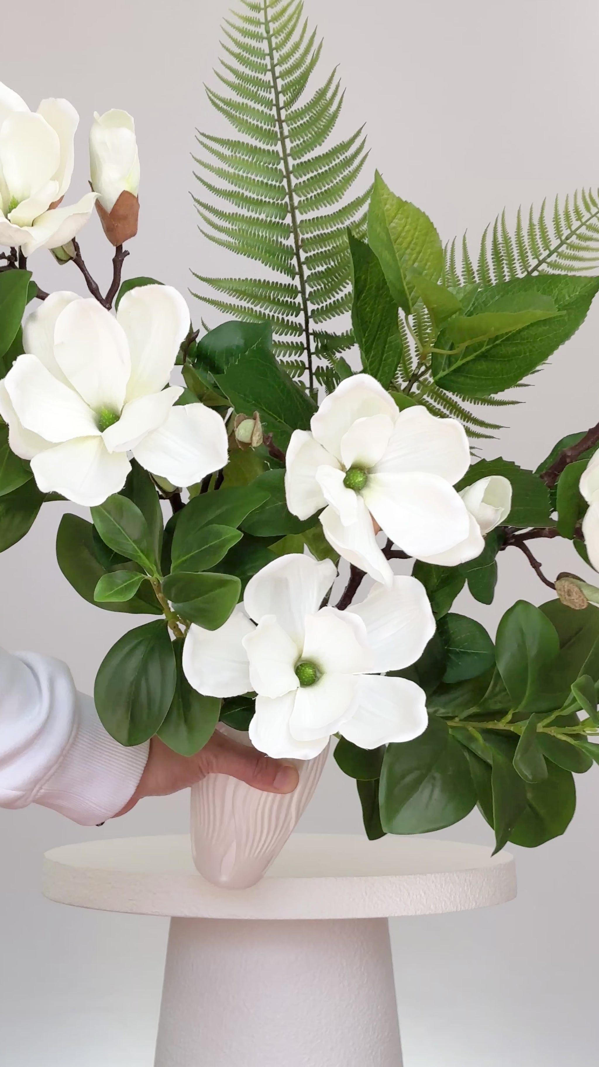 Artificial white magnolia flower arrangement with lush green leaves and fern accents, beautifully styled in a sleek white vase on a round white table, set against a neutral background.