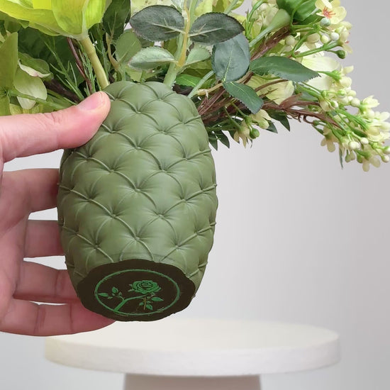 Artificial floral arrangement featuring cream roses, green orchids, and lush ferns in a textured olive-green vase, styled on a white pedestal — perfect for adding a refreshing, natural touch to modern or earthy-toned interiors.