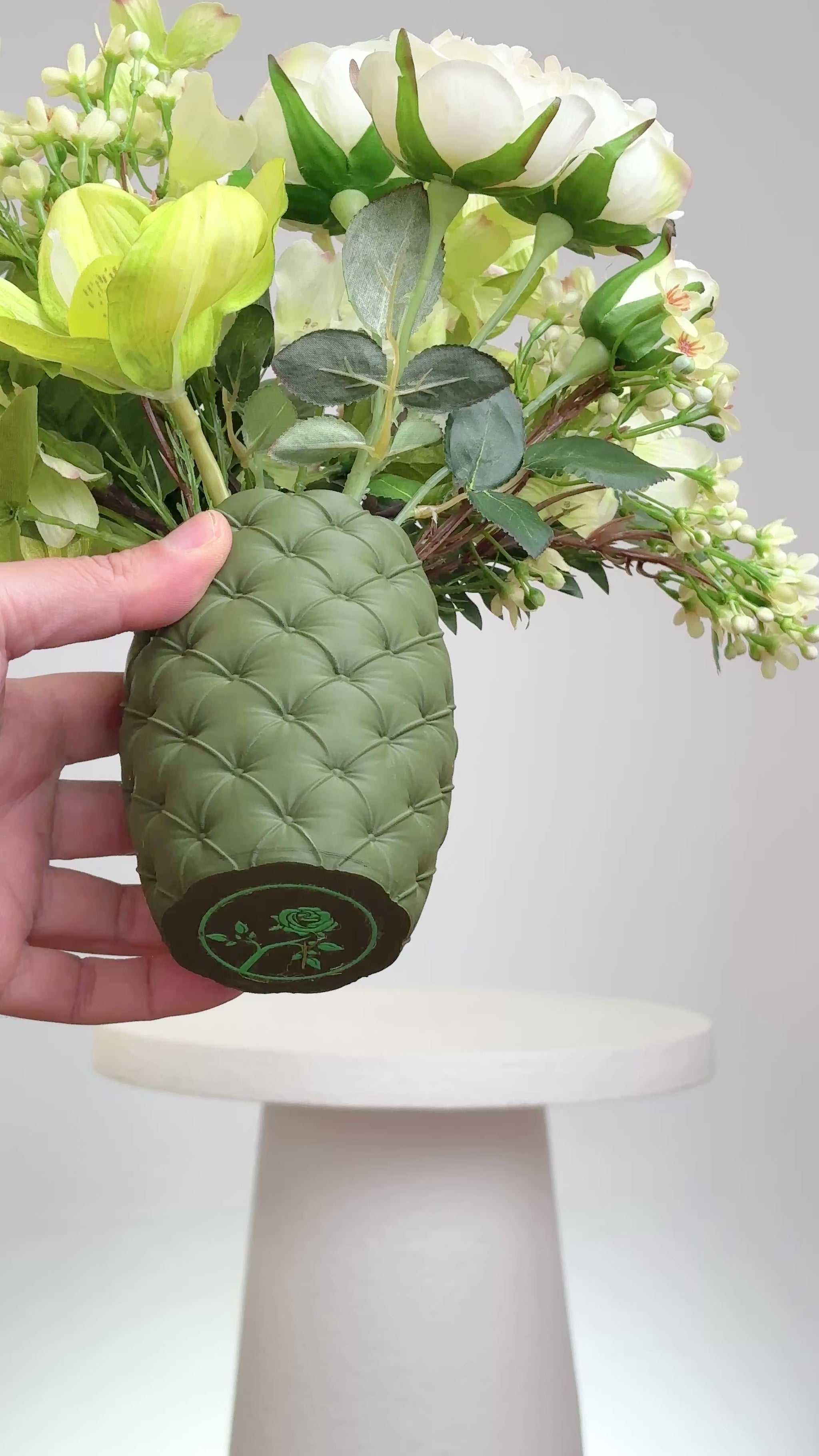 Artificial floral arrangement featuring cream roses, green orchids, and lush ferns in a textured olive-green vase, styled on a white pedestal — perfect for adding a refreshing, natural touch to modern or earthy-toned interiors.