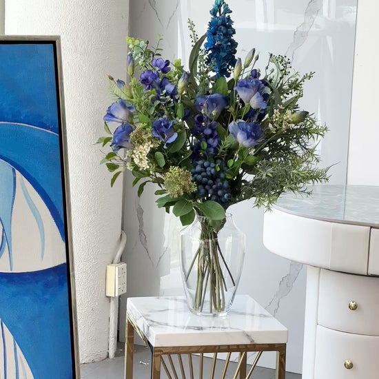 Artificial blue floral arrangement with greenery and berry accents, displayed in a clear glass vase with acrylic water