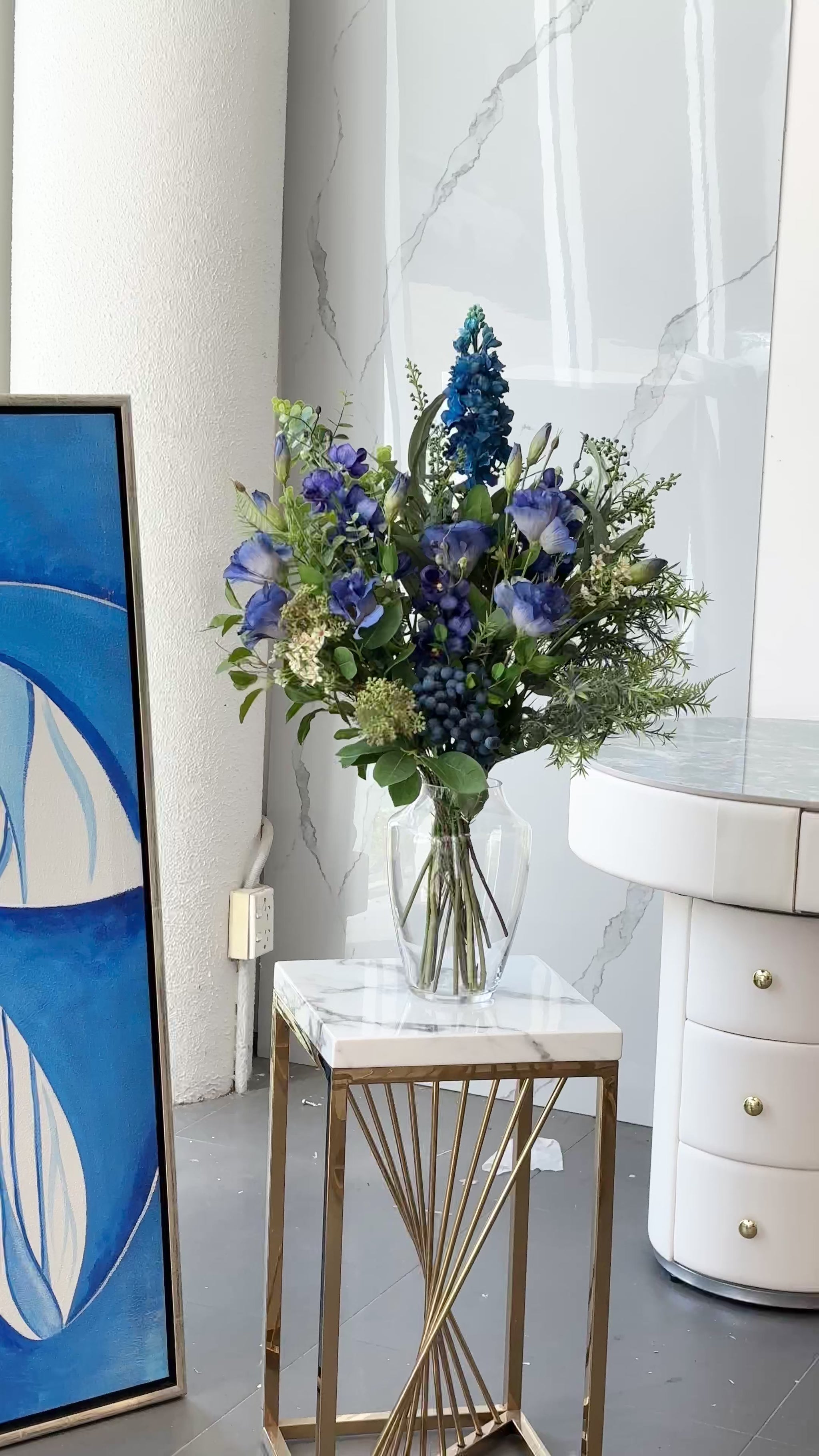 Artificial blue floral arrangement with greenery and berry accents, displayed in a clear glass vase with acrylic water