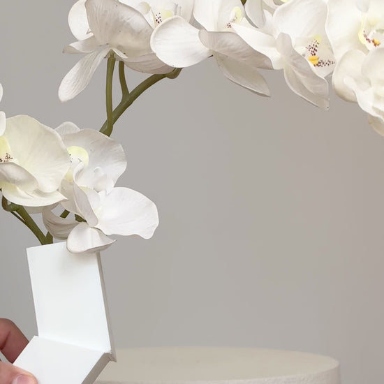 Artificial white orchid arrangement cascading gracefully from a modern geometric white vase, displayed on a white pedestal against a minimalist neutral background.