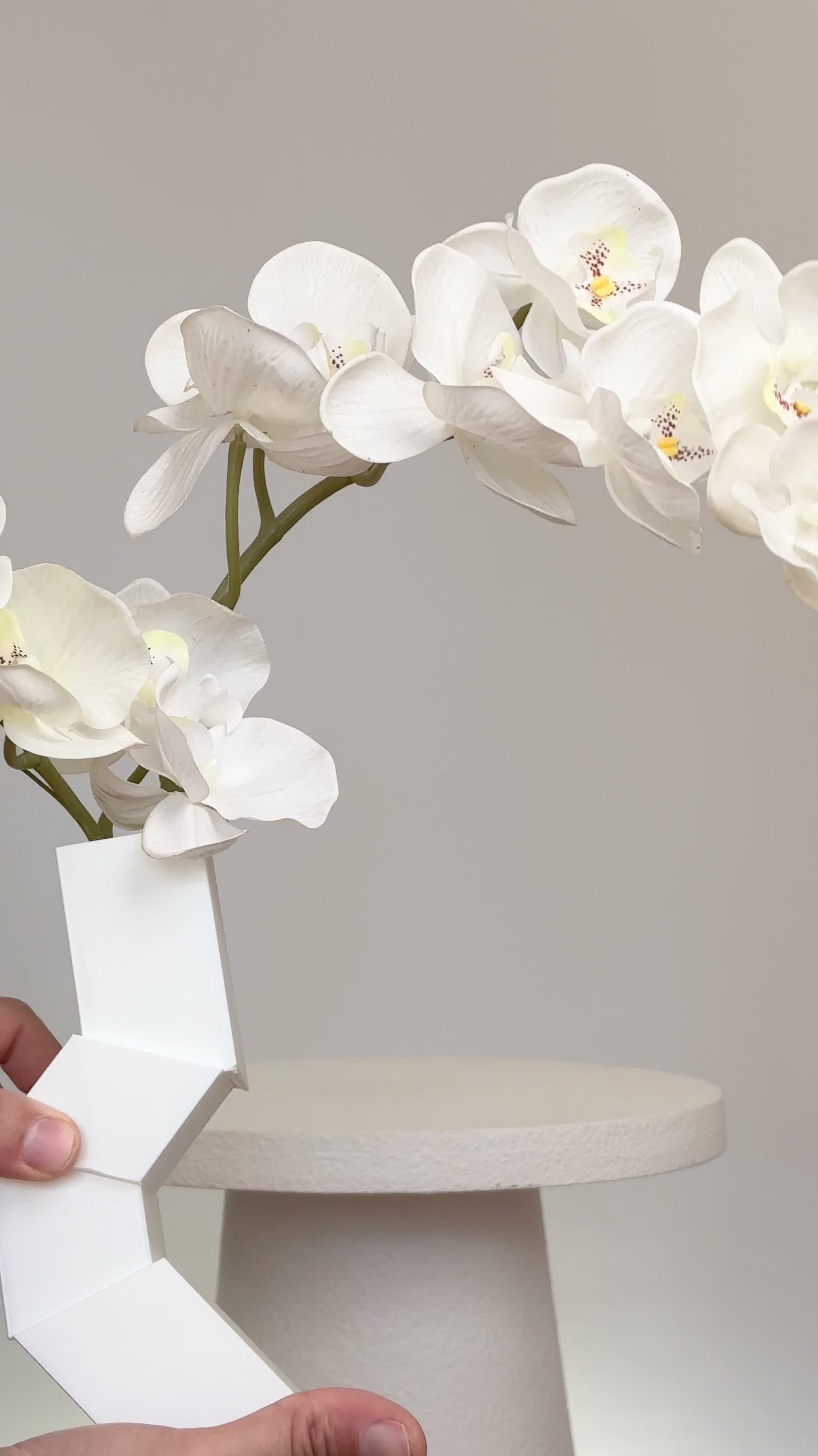 Artificial white orchid arrangement cascading gracefully from a modern geometric white vase, displayed on a white pedestal against a minimalist neutral background.