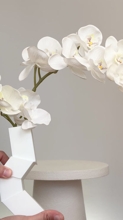 Artificial white orchid arrangement cascading gracefully from a modern geometric white vase, displayed on a white pedestal against a minimalist neutral background.