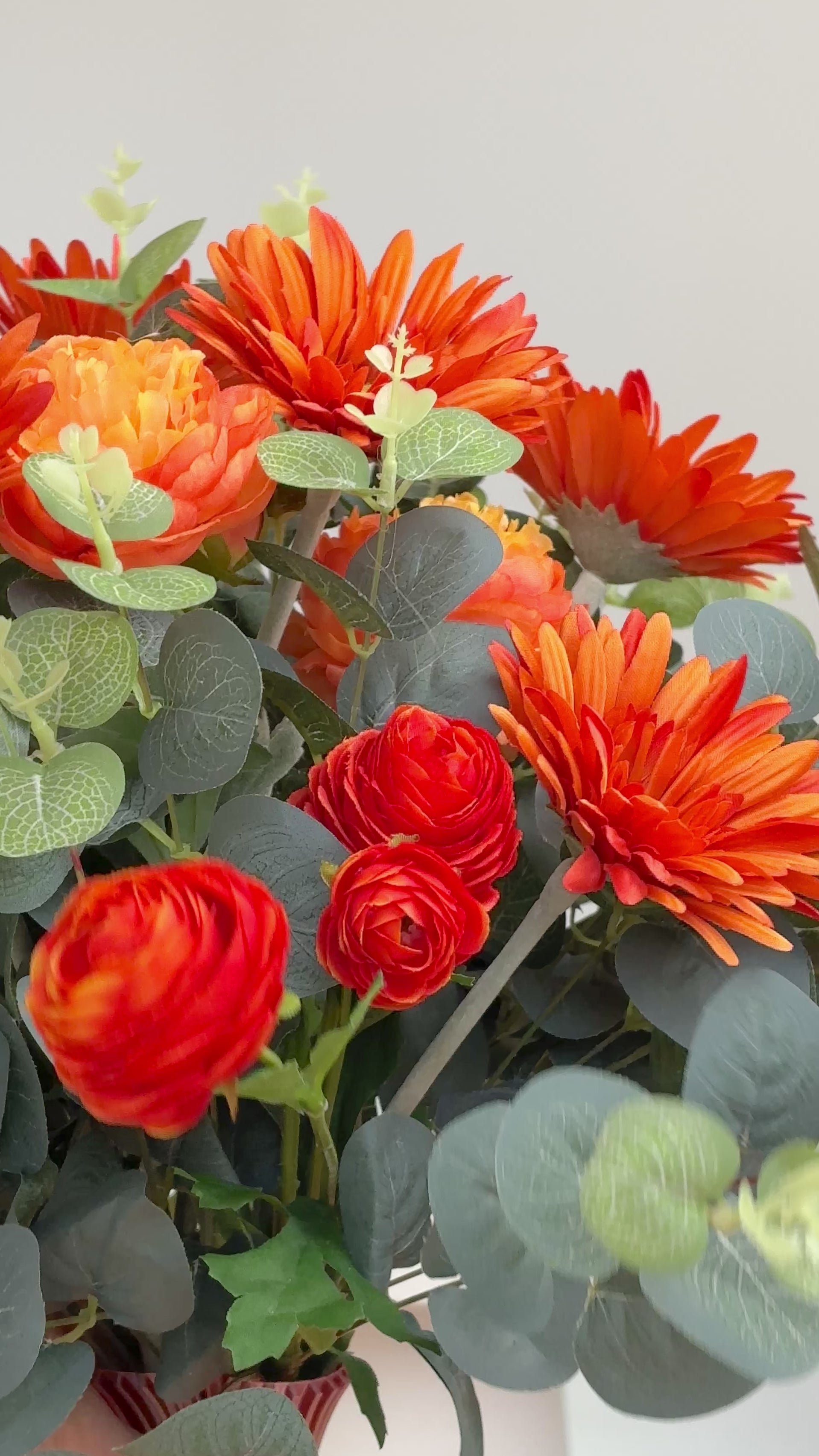 Artificial floral arrangement with orange gerberas and ranunculus surrounded by eucalyptus leaves, displayed in an ombre vase blending amber and green tones on a light background.
