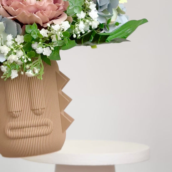 Artificial arrangement with lush ferns, blush peonies, hydrangeas, and white baby’s breath in a terracotta sculptural vase, styled on a marble pedestal against a light background.
