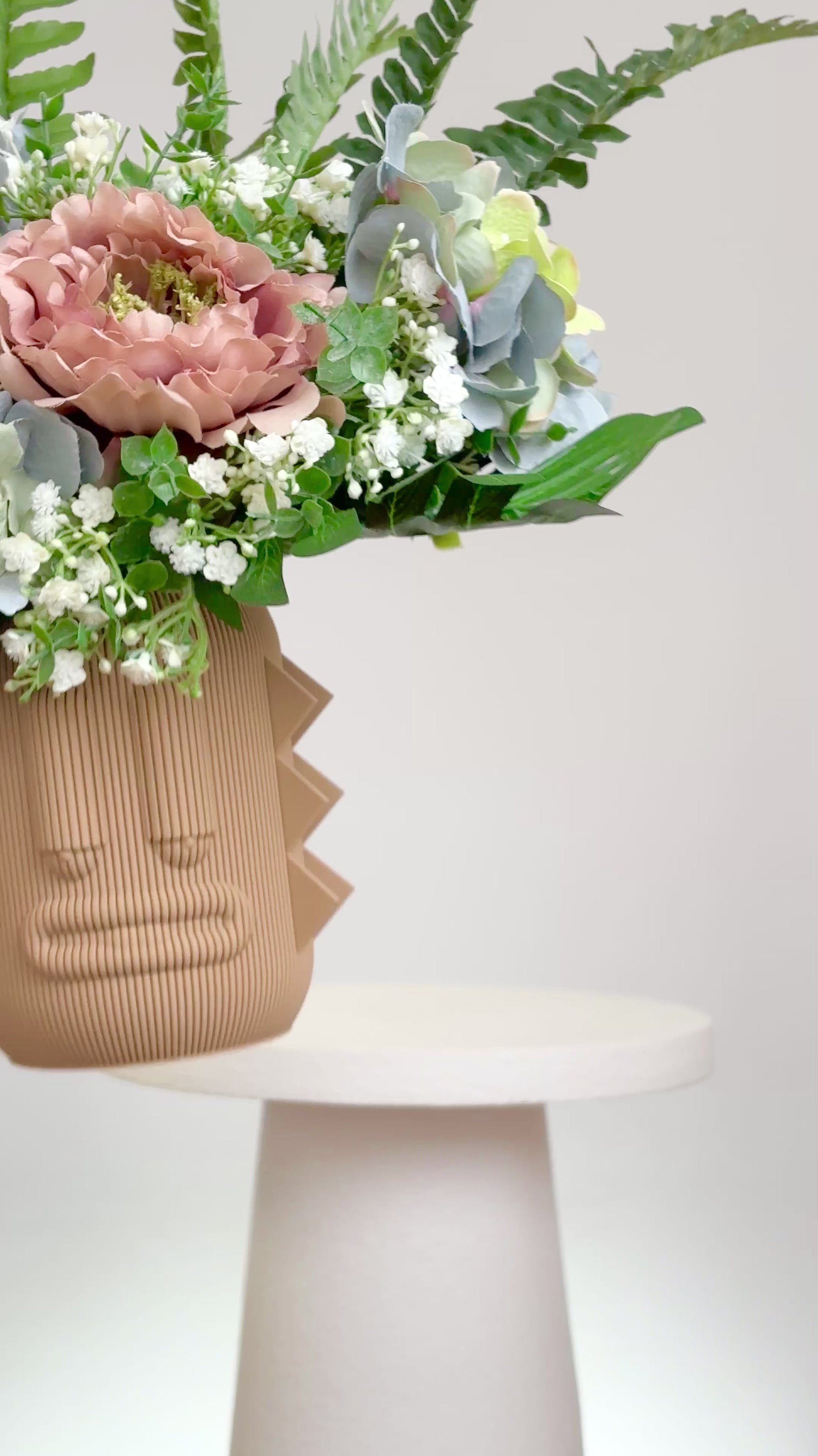 Artificial arrangement with lush ferns, blush peonies, hydrangeas, and white baby’s breath in a terracotta sculptural vase, styled on a marble pedestal against a light background.