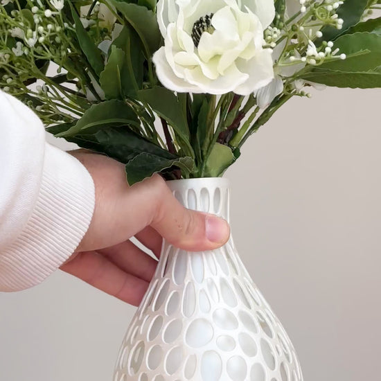 Artificial floral arrangement featuring ivory roses, white lisianthus, and blossoms with lush green foliage, styled in a white honeycomb-textured vase on a neutral background.