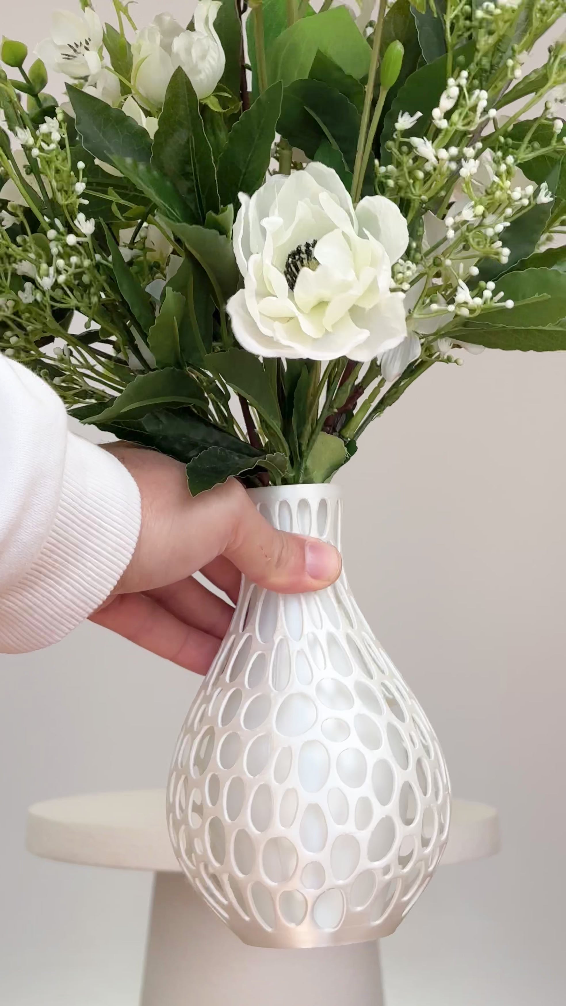Artificial floral arrangement featuring ivory roses, white lisianthus, and blossoms with lush green foliage, styled in a white honeycomb-textured vase on a neutral background.