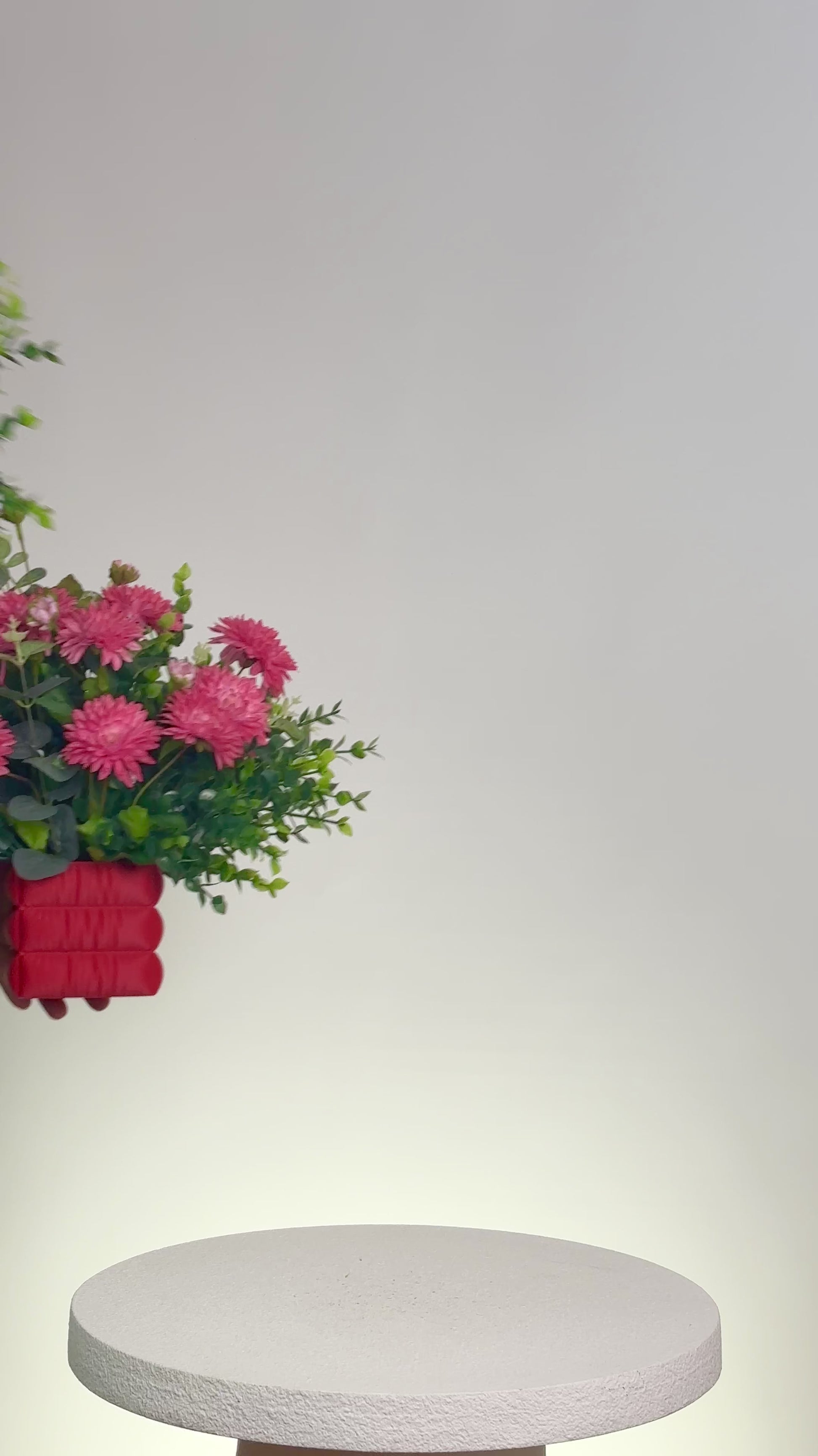 Artificial floral arrangement featuring bright pink chrysanthemums and green foliage in a soft red padded square vase, styled on a white pedestal — perfect for adding a cheerful pop of color to modern interiors.