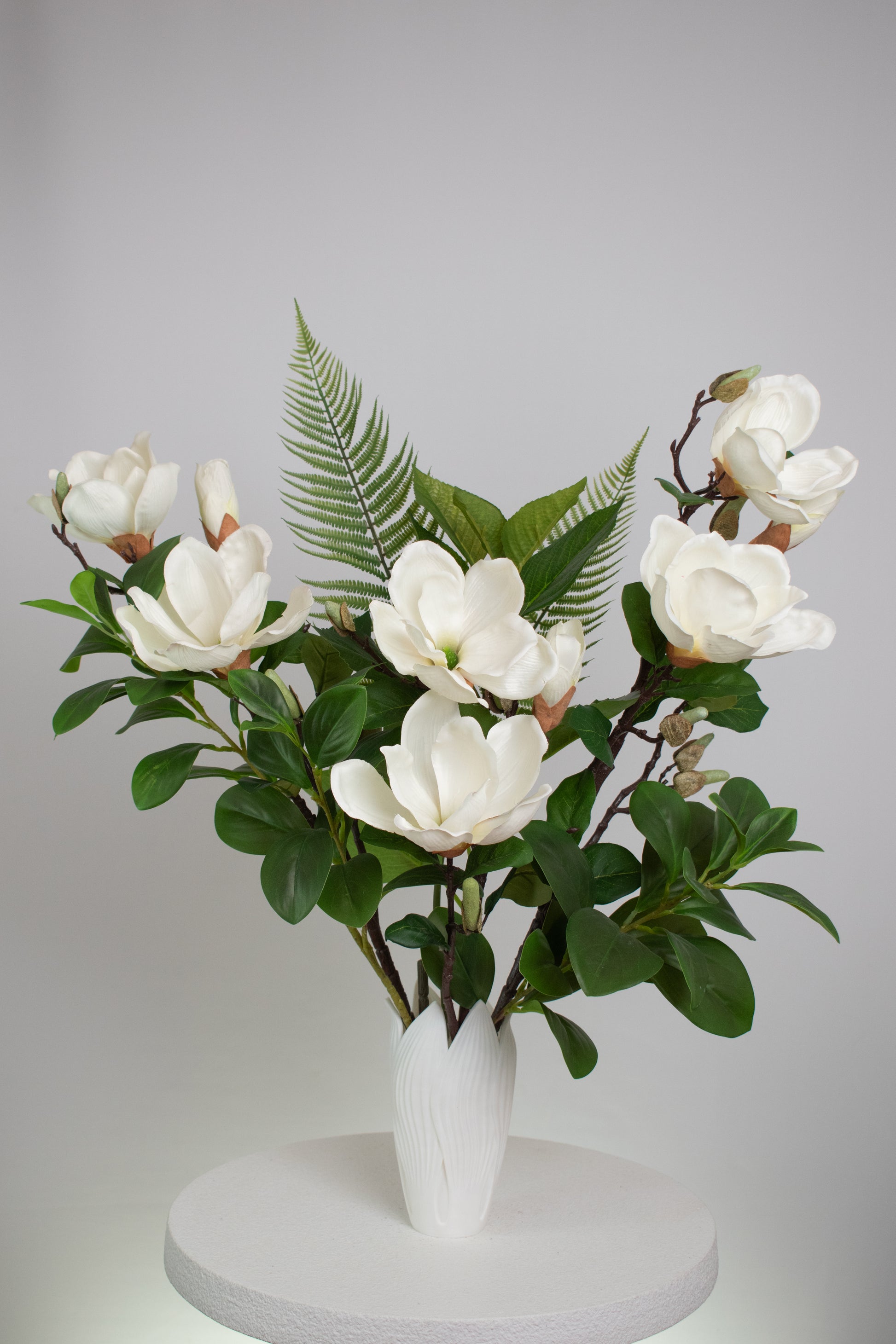 Artificial white magnolia flower arrangement with lush green leaves and fern accents, beautifully styled in a sleek white vase on a round white table, set against a neutral background