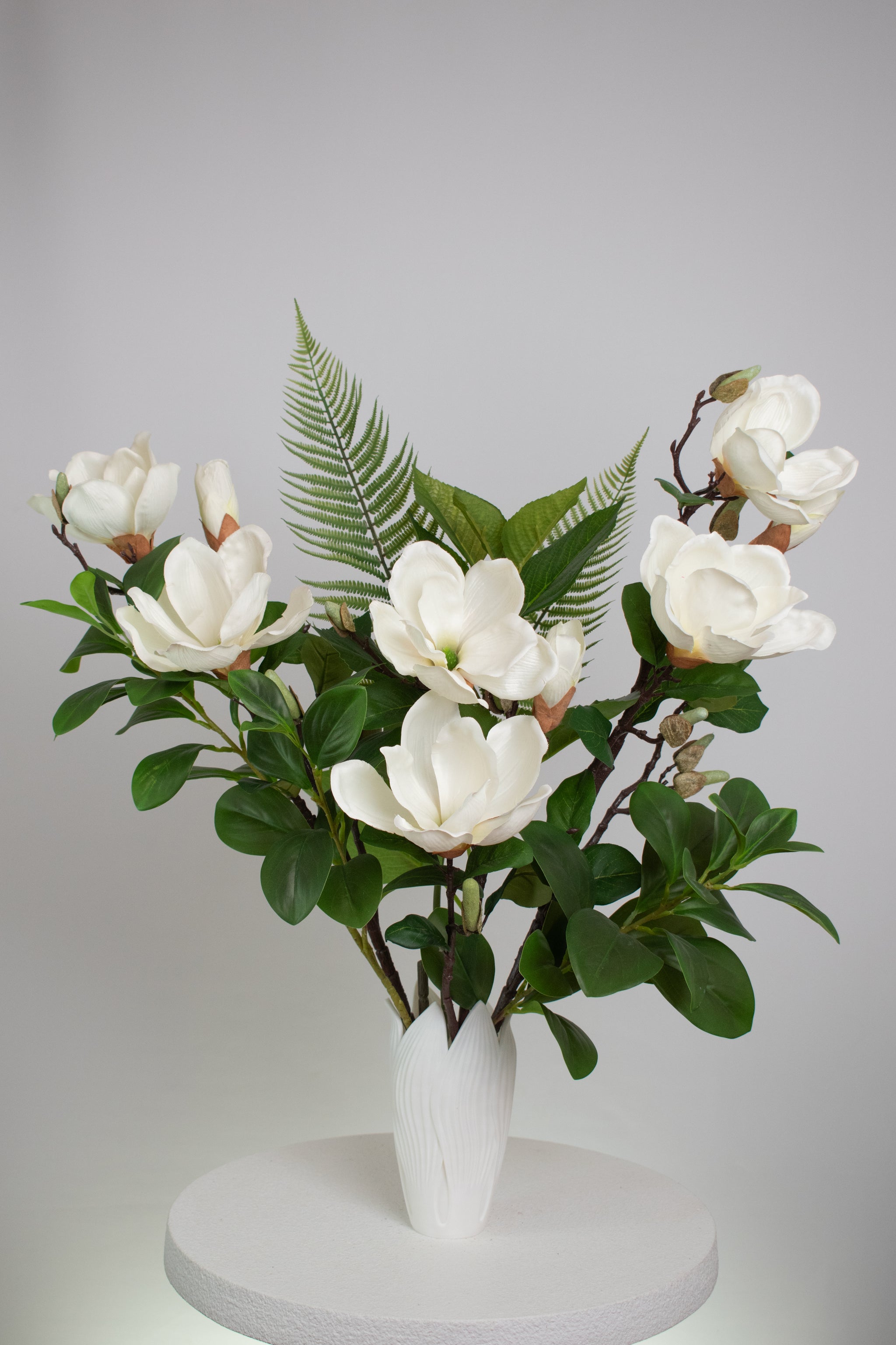 Artificial white magnolia flower arrangement with lush green leaves and fern accents, beautifully styled in a sleek white vase on a round white table, set against a neutral background