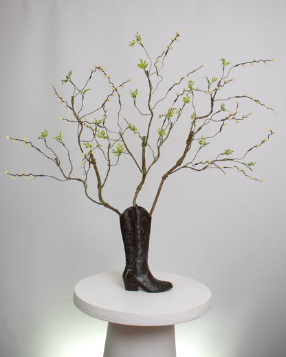 Rustic artificial floral arrangement with natural branches and light green leaves arranged in a dark brown cowboy boot-shaped vase, styled as a modern rustic décor accent.