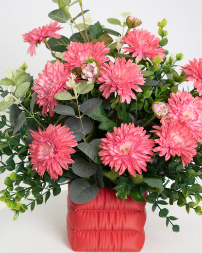 Artificial floral arrangement featuring bright pink chrysanthemums and green foliage in a soft red padded square vase, styled on a white pedestal — perfect for adding a cheerful pop of color to modern interiors.