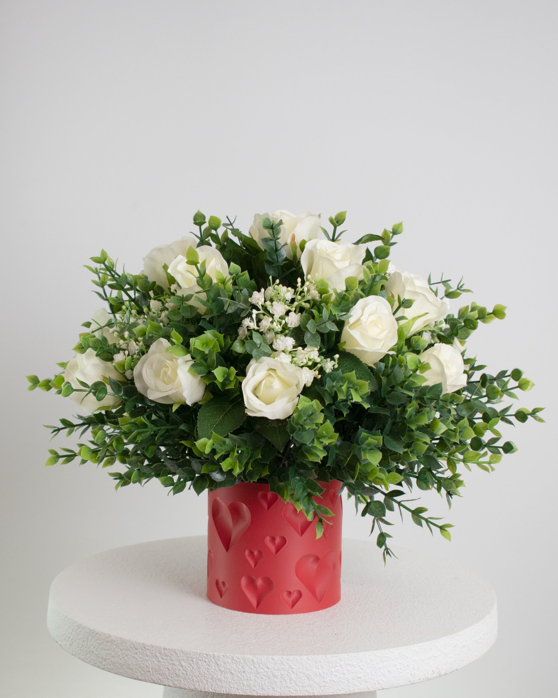 A romantic artificial flower arrangement featuring elegant white roses and lush green foliage displayed in a red vase embossed with heart shapes, symbolizing love and affection.