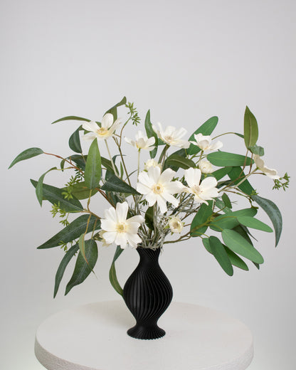 Artificial arrangement featuring white daisy-like flowers and lush eucalyptus leaves in a sculpted matte black vase, styled on a white pedestal — perfect for modern, minimalist, or monochrome interiors.