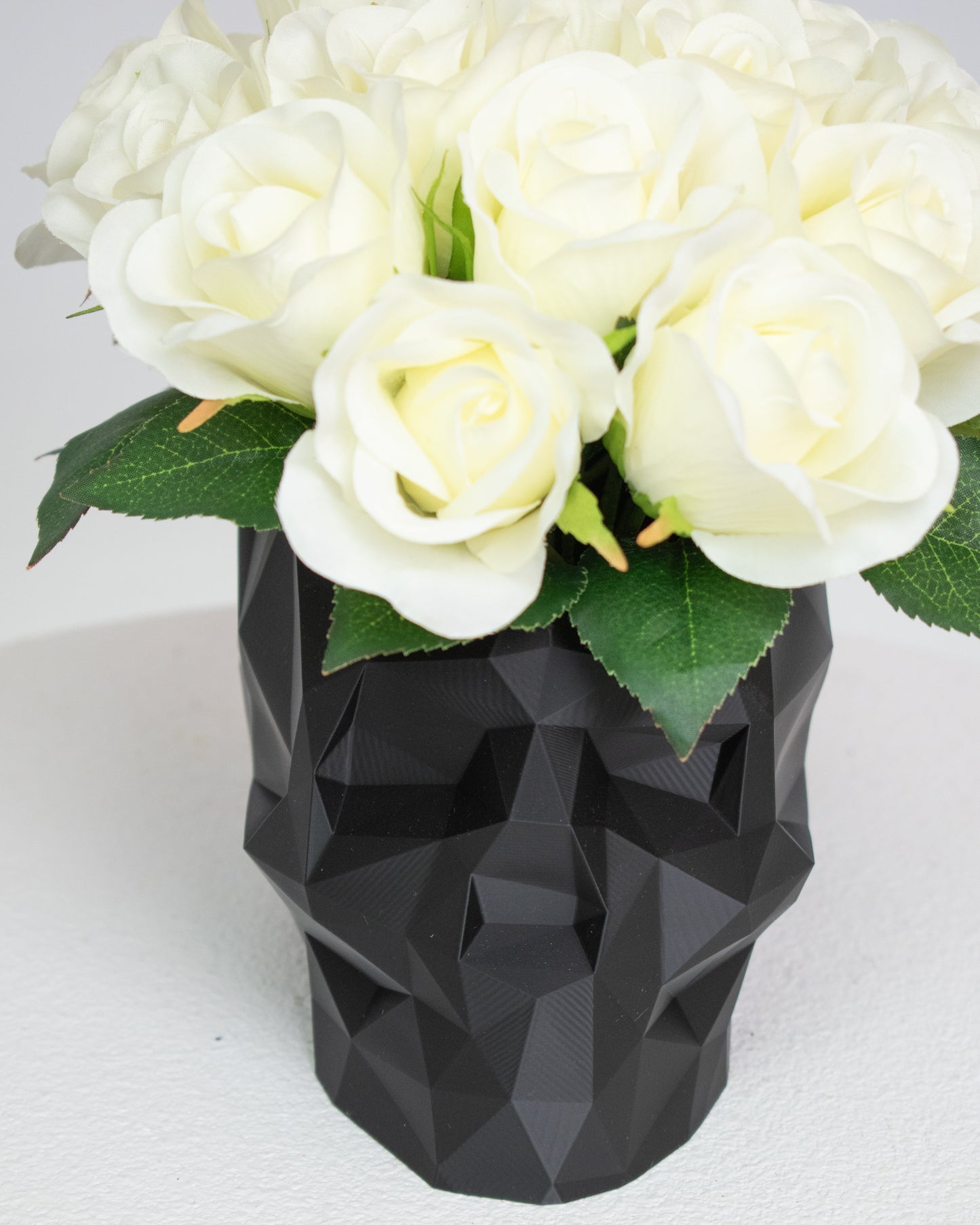 Artificial ivory rose arrangement displayed in a black geometric skull vase on a white pedestal against a neutral background.