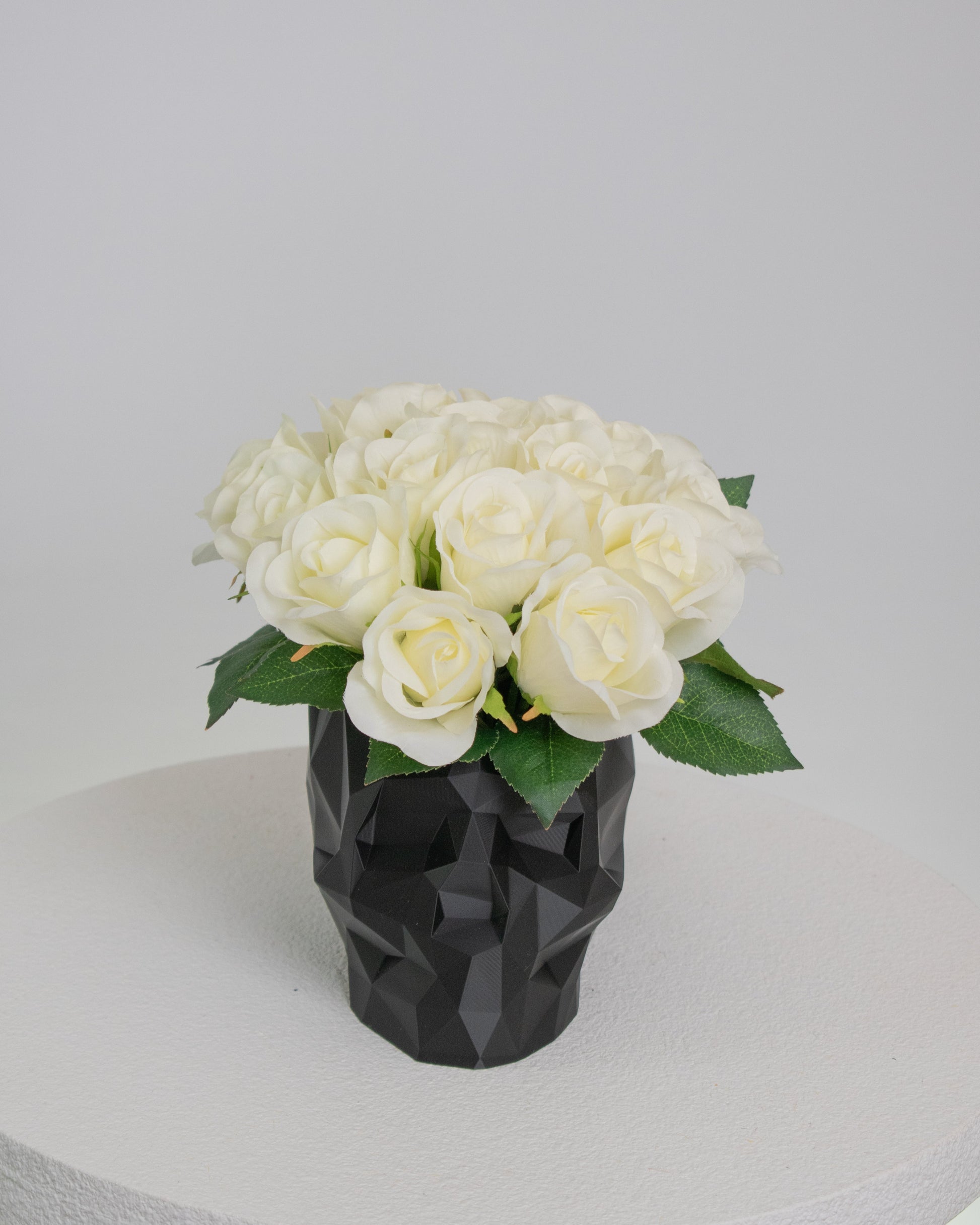 Artificial ivory rose arrangement displayed in a black geometric skull vase on a white pedestal against a neutral background.