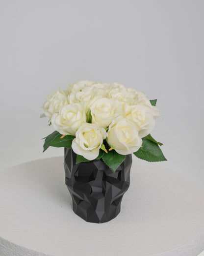 Artificial ivory rose arrangement displayed in a black geometric skull vase on a white pedestal against a neutral background.