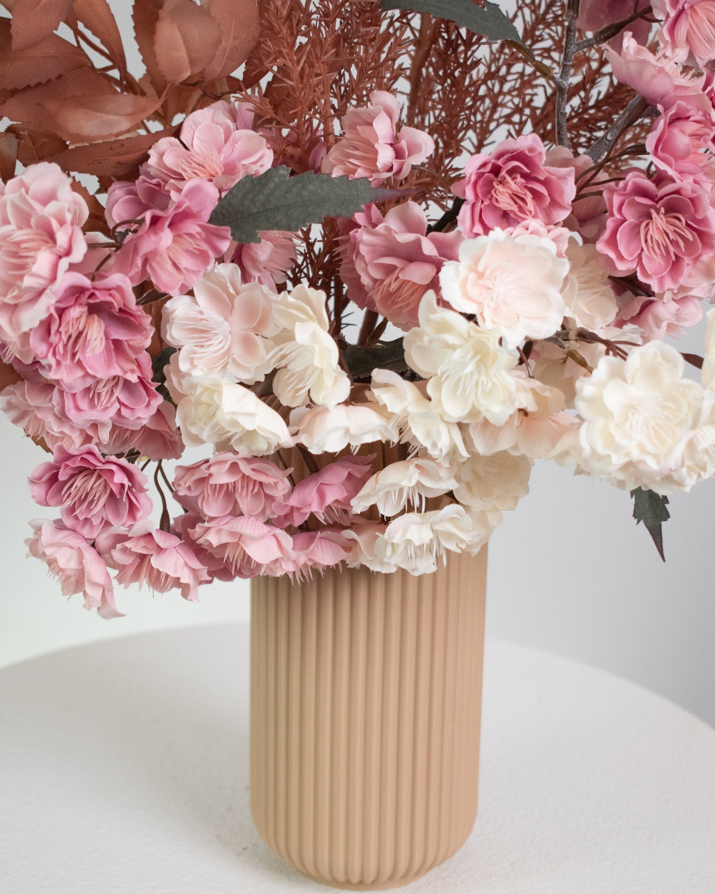 Artificial floral arrangement featuring pink and ivory blossoms with rust-colored foliage, elegantly styled in a tall ribbed nude vase, displayed on a white round pedestal against a neutral background.