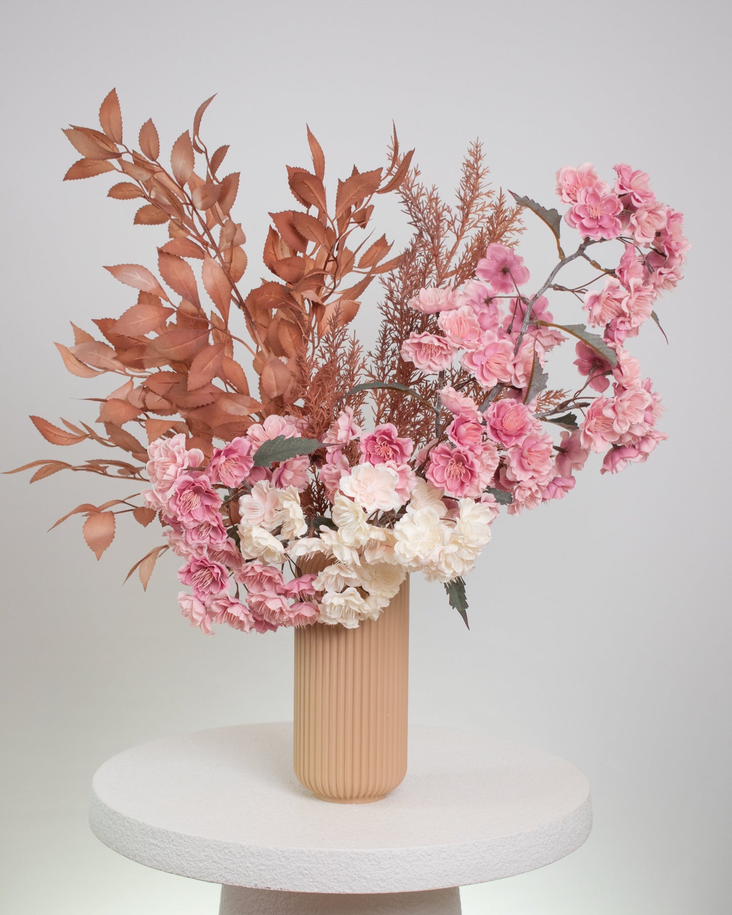 Artificial floral arrangement featuring pink and ivory blossoms with rust-colored foliage, elegantly styled in a tall ribbed nude vase, displayed on a white round pedestal against a neutral background.