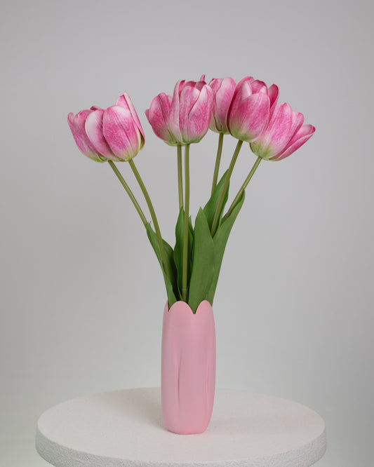 Artificial pink tulip arrangement with lifelike green stems and leaves, displayed in a matching pink vase with a scalloped tulip-shaped rim, set against a neutral background.