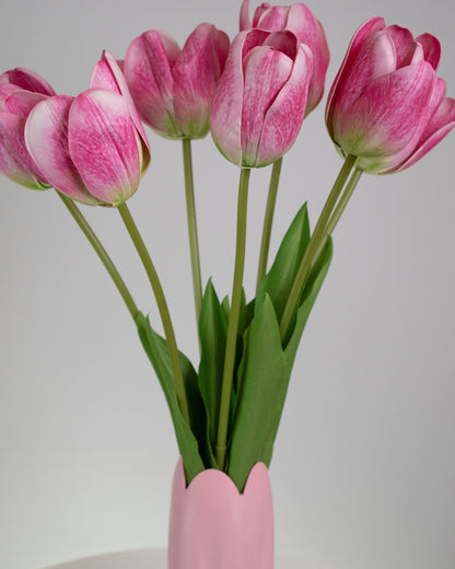 Artificial pink tulip arrangement with lifelike green stems and leaves, displayed in a matching pink vase with a scalloped tulip-shaped rim, set against a neutral background.
