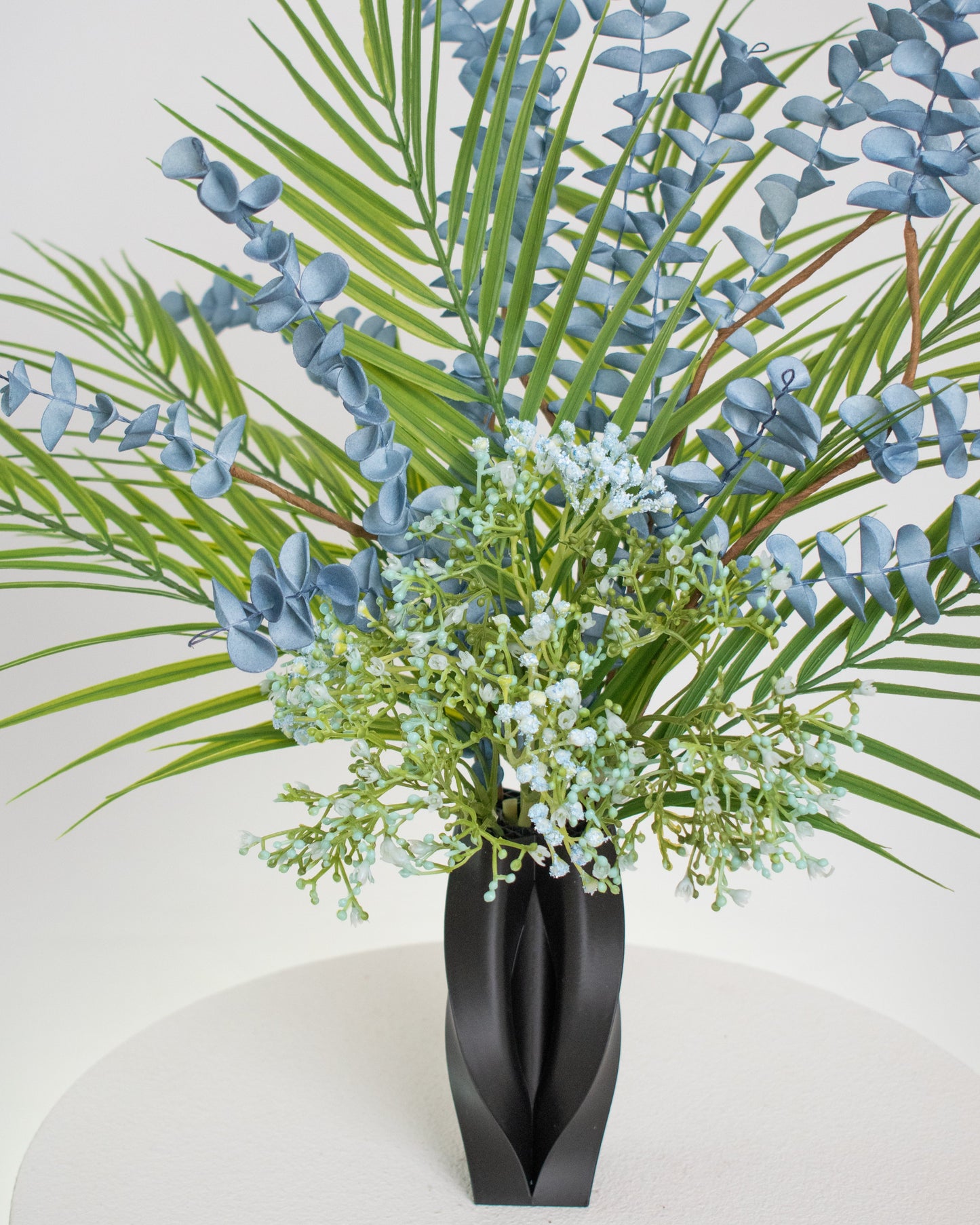 Artificial arrangement with blue eucalyptus stems, palm leaves, and delicate greenery in a sleek black sculpted vase, styled on a white pedestal — ideal for modern or minimalist interiors with cool or neutral tones.