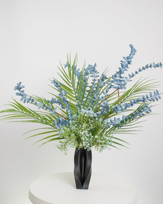 Artificial arrangement with blue eucalyptus stems, palm leaves, and delicate greenery in a sleek black sculpted vase, styled on a white pedestal — ideal for modern or minimalist interiors with cool or neutral tones.
