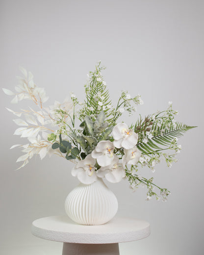 Artificial white orchid floral arrangement in a ribbed white vase with lush greenery and baby’s breath.