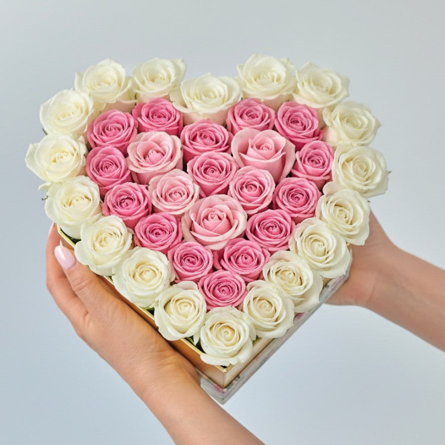 Heart-shaped arrangement of pink and white roses held by a person against a light gray background