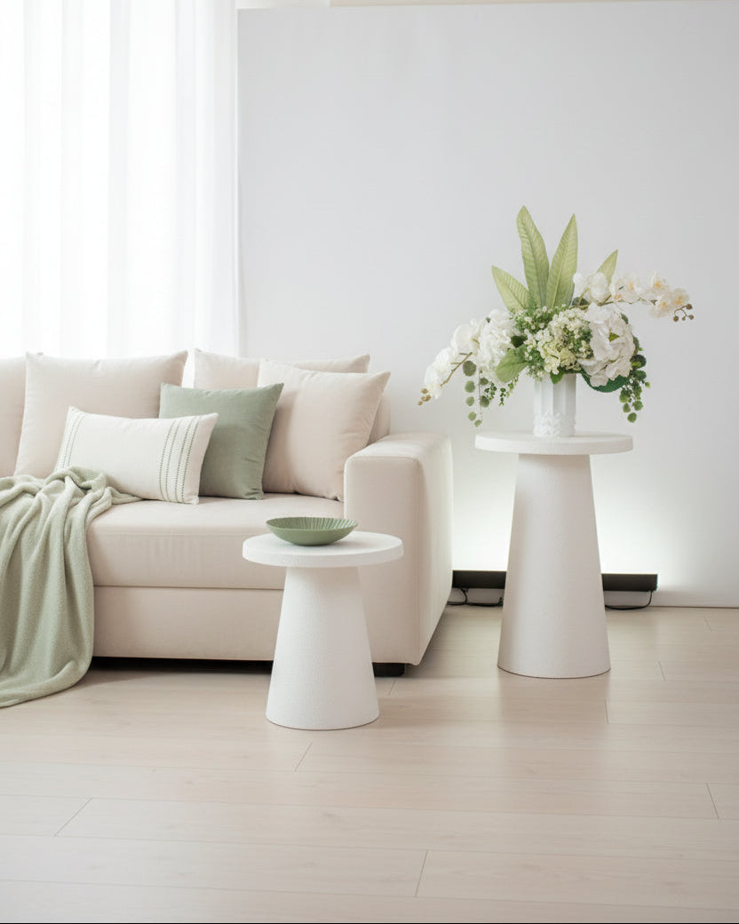 Artificial white floral arrangement featuring cascading orchids, hydrangeas, and lush greenery with tall tropical leaves, beautifully displayed in a textured white ceramic vase on a round white pedestal against a light background.