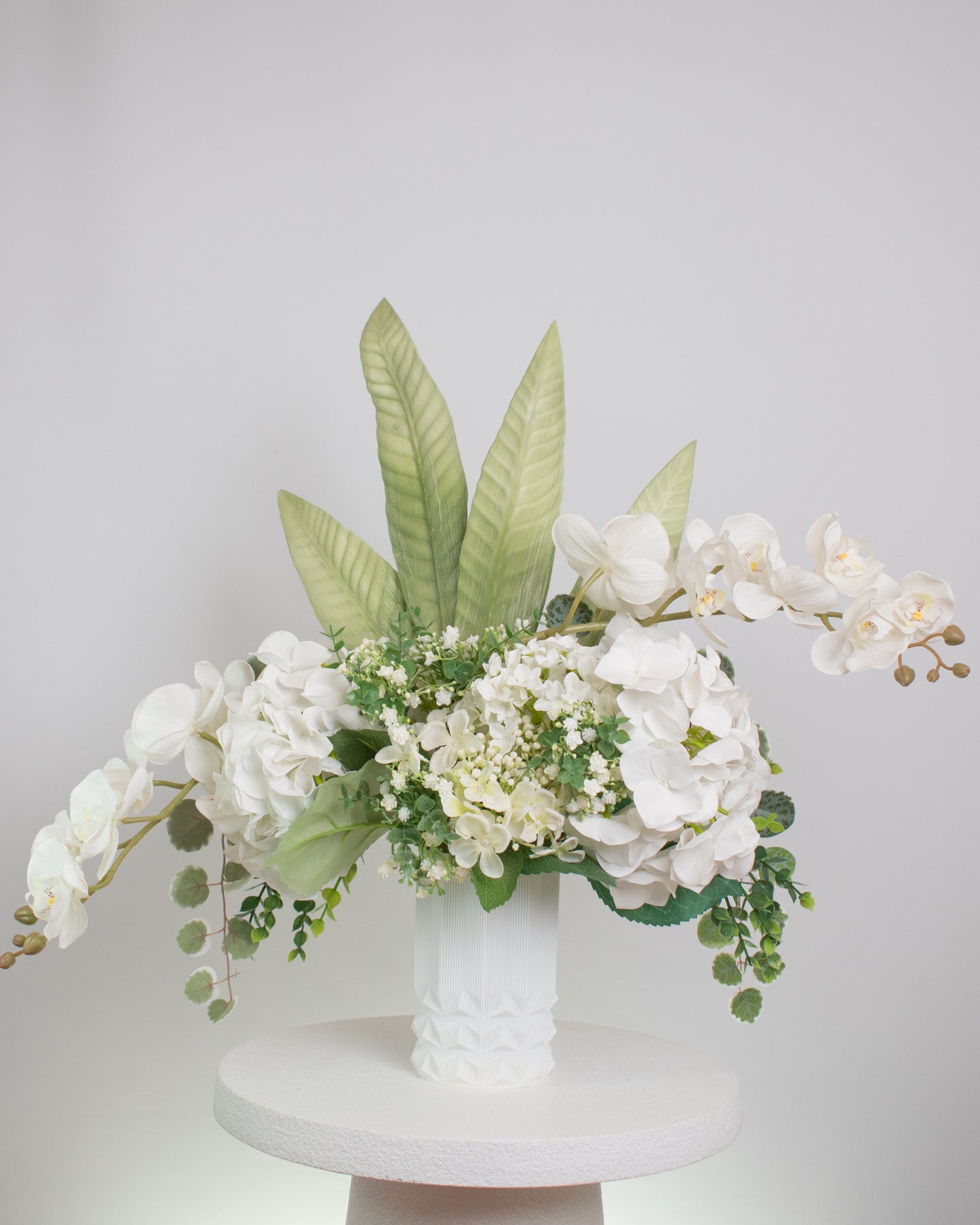 Artificial white floral arrangement featuring cascading orchids, hydrangeas, and lush greenery with tall tropical leaves, beautifully displayed in a textured white ceramic vase on a round white pedestal against a light background.