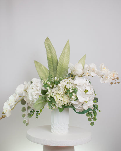 Artificial white floral arrangement featuring cascading orchids, hydrangeas, and lush greenery with tall tropical leaves, beautifully displayed in a textured white ceramic vase on a round white pedestal against a light background.