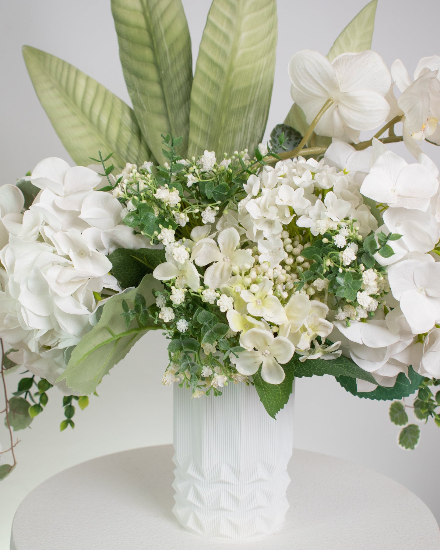Artificial white floral arrangement featuring cascading orchids, hydrangeas, and lush greenery with tall tropical leaves, beautifully displayed in a textured white ceramic vase on a round white pedestal against a light background.