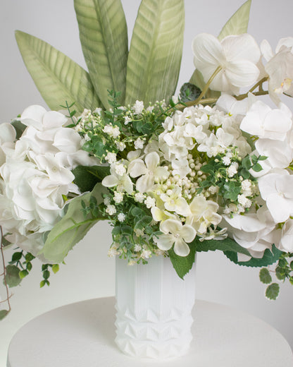 Artificial white floral arrangement featuring cascading orchids, hydrangeas, and lush greenery with tall tropical leaves, beautifully displayed in a textured white ceramic vase on a round white pedestal against a light background.