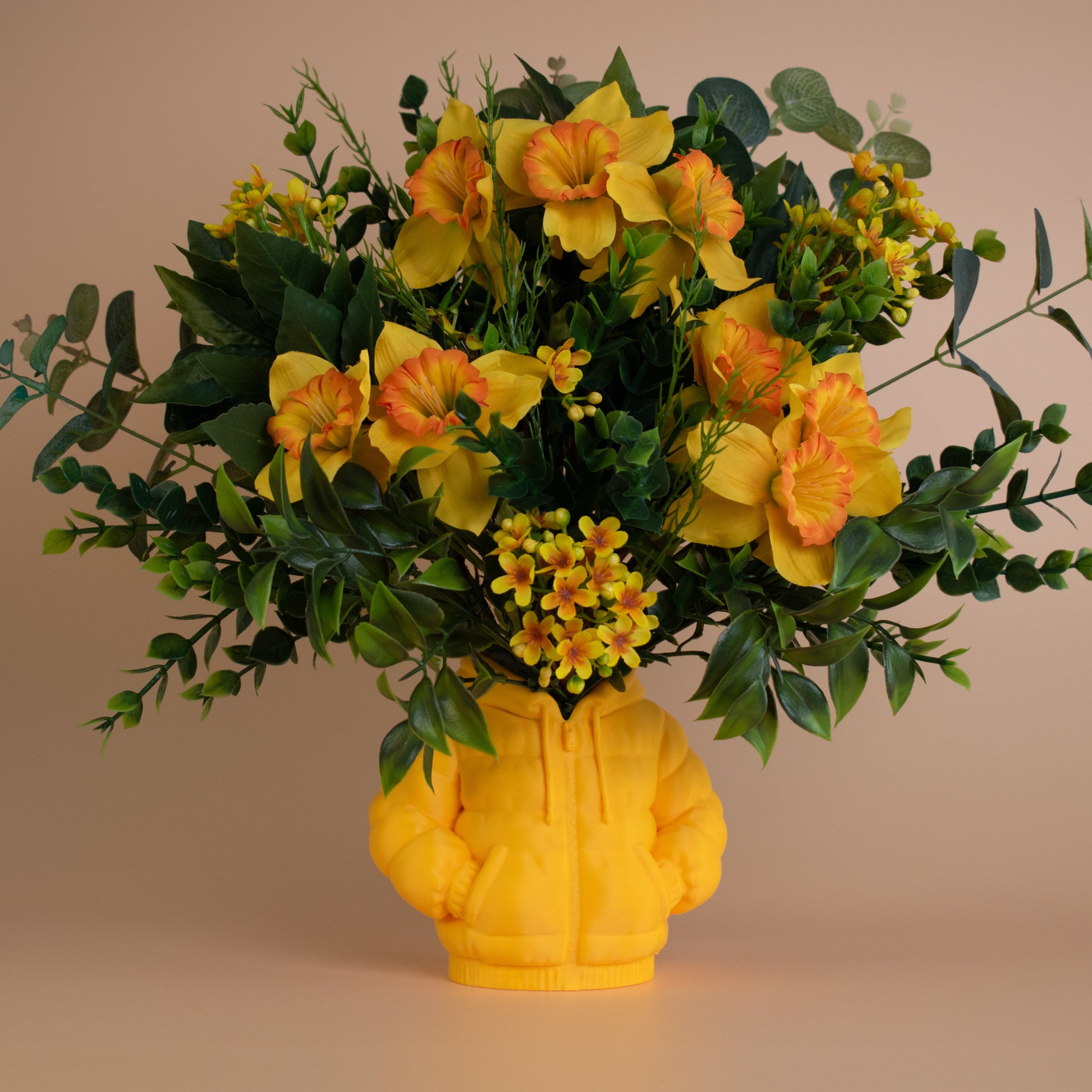 Bright yellow and orange artificial flower arrangement in a hoodie-shaped yellow vase, surrounded by lush green leaves on a neutral background.