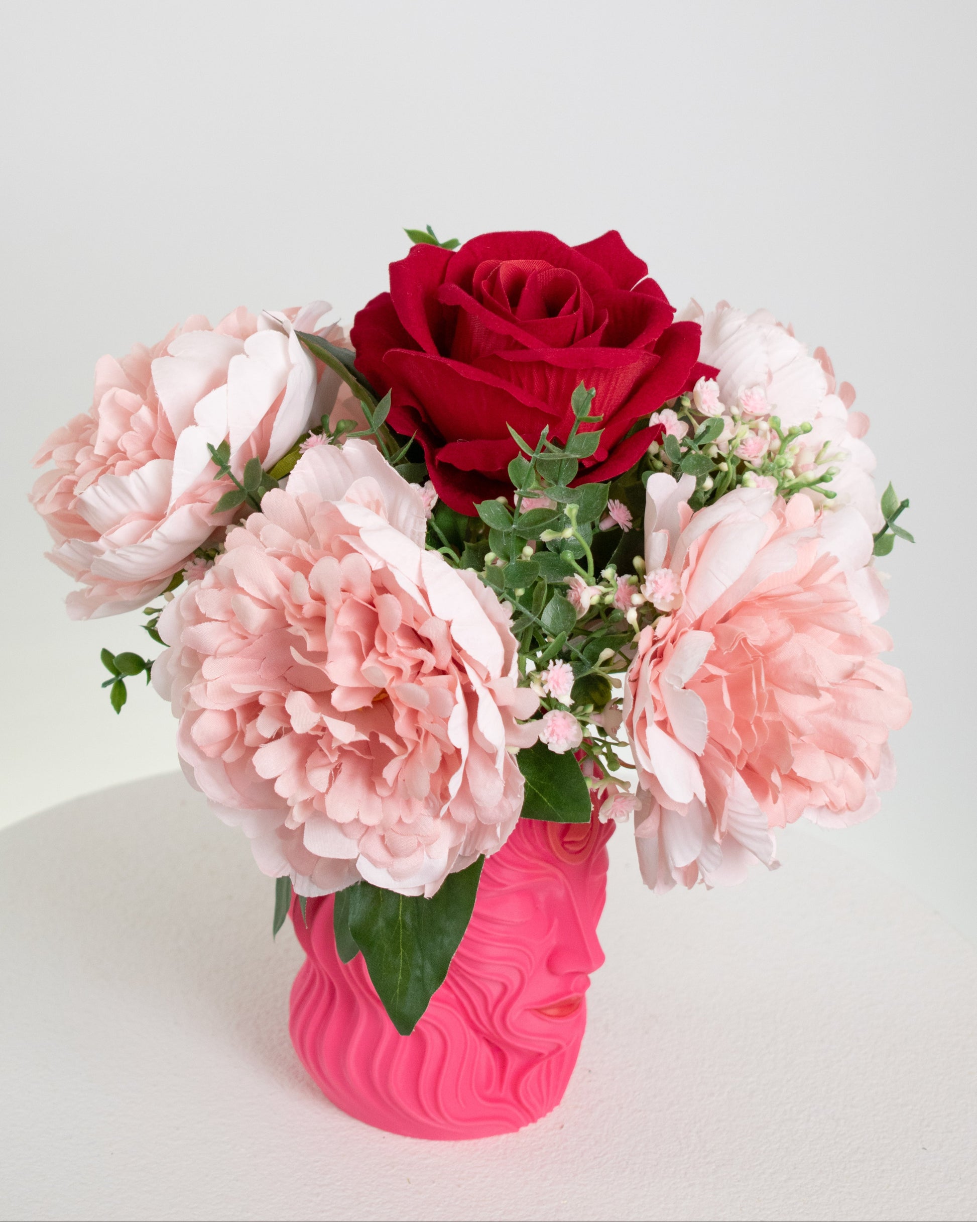 Artificial floral arrangement with blush pink peonies and a red rose in a sculpted pink vase shaped like a woman’s face, styled on a white pedestal — elegant and feminine décor for modern interiors.